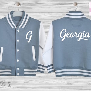 May include: A blue and white varsity jacket featuring a white letter G on the front left chest and the name Georgia in white script lettering on the back. The jacket has white sleeves, a striped collar, cuffs, and hem, and white snap buttons.