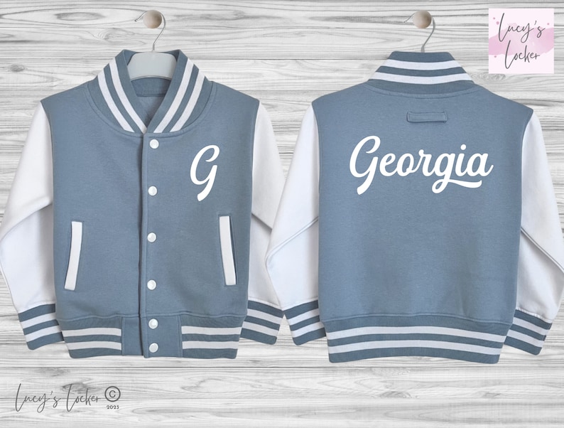 May include: A blue and white varsity jacket featuring a white letter G on the front left chest and the name Georgia in white script lettering on the back. The jacket has white sleeves, a striped collar, cuffs, and hem, and white snap buttons.