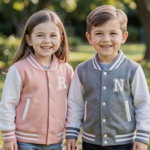 May include: Two children wearing letterman jackets. One jacket is dusty rose with white sleeves and the letter "R". The other jacket is grey with white sleeves and the letter "N". Both jackets have white and grey striped trim.