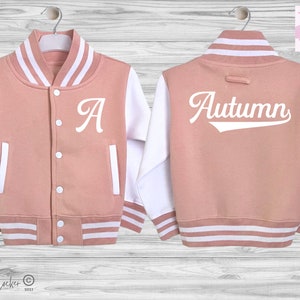 May include: A dusty rose varsity jacket with white sleeves, featuring a white letter A on the front left chest and the name Autumn in white script on the back. The jacket includes a snap-button front, white striped ribbed trim, and pockets.