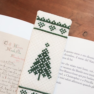 Pode incluir: Marcador de livros artesanal com um desenho de árvore de Natal em ponto cruz verde escuro sobre um fundo creme. O marcador tem bordas decorativas e está colocado em um livro aberto com texto manuscrito e um pinha.