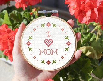 Puede incluir: Un aro de bordado blanco con un diseño de punto de cruz que dice "Te amo mamá" con un corazón rosa. El aro está rodeado de una corona de flores rosas con hojas verdes.