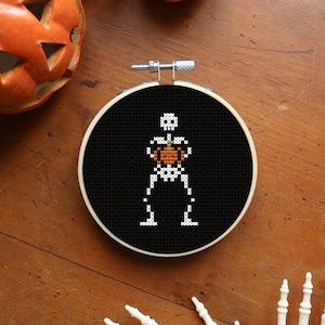 May include: A black embroidery hoop featuring a white skeleton holding a small orange pumpkin. The hoop is on a wooden surface with two orange pumpkins and a lit candle.