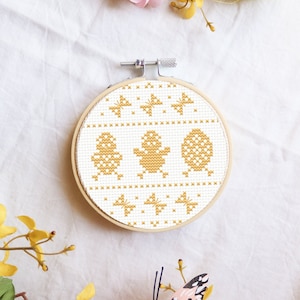 May include: A white embroidery hoop with a cross-stitch design of three yellow chicks. The design is on a white background and the hoop is made of wood.