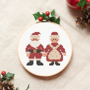 May include: Cross-stitch embroidery hoop with a white background featuring a red and white Santa Claus and Mrs. Claus holding hands. The embroidery is framed in a wooden hoop.