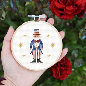 May include: A white embroidery hoop with a cross-stitch design of Uncle Sam. The design features a blue jacket, red and white striped pants, a white hat with a red, white, and blue star design, and a white beard. The background is white with gold stars.