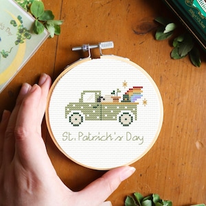 May include: A St. Patrick's Day cross-stitch embroidery hoop featuring a green truck carrying a pot of gold and a rainbow. The text "St. Patrick's Day" is below the truck. Vintage postcards and a green tin are in the background.