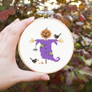 Peut inclure: Un épouvantail à tête de citrouille orange et violette avec un patch cousu et deux corbeaux noirs. L'épouvantail tient une râteau et se tient sur un fond blanc.