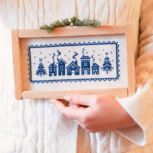 May include: A wooden frame with a white cross-stitch design of a winter village scene. The design features blue houses with white snow and trees.