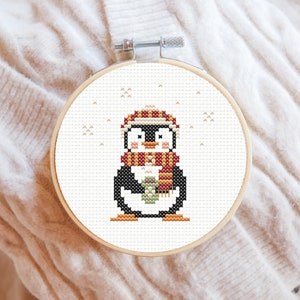May include: A cross-stitch embroidery hoop with a white background and a penguin wearing a red and yellow scarf and hat. The penguin is holding a green ball. The background is white with small white snowflakes.