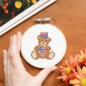 May include: A cross-stitch embroidery hoop featuring a brown teddy bear wearing a patriotic hat and a flag-themed vest. The bear is stitched on white fabric, held in a wooden hoop. The image includes flowers and a magazine.