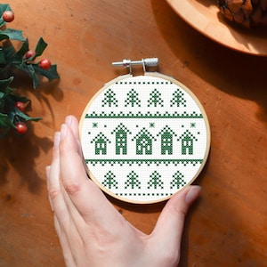 May include: A round cross-stitch embroidery hoop featuring a festive green and white design. The pattern includes rows of Christmas trees, houses, and stars. The embroidery is set in a wooden hoop, perfect for holiday decor.