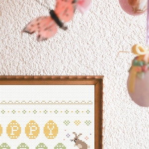 May include: A framed cross-stitch design featuring a bunny rabbit surrounded by yellow and green Easter eggs. The design is on white fabric with a brown wooden frame.