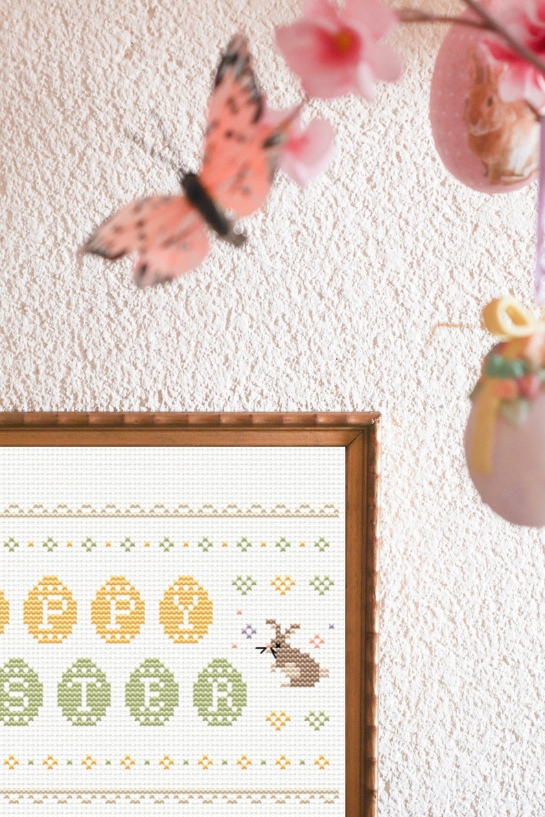 May include: A framed cross-stitch design featuring a bunny rabbit surrounded by yellow and green Easter eggs. The design is on white fabric with a brown wooden frame.