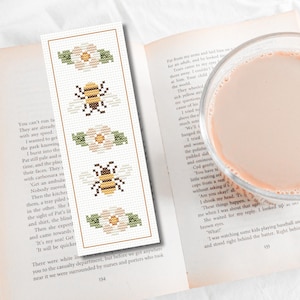 Può includere: Un segnalibro fatto a mano con un disegno a punto croce con api e fiori. Il segnalibro è rettangolare con uno sfondo color crema e un bordo marrone. Il design include due api e due fiori.