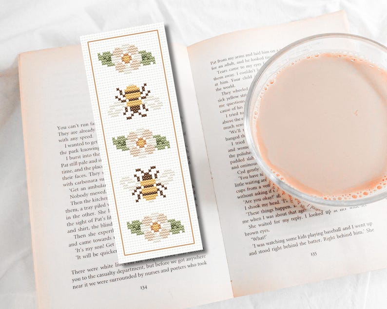 May include: A handmade bookmark with a cross-stitch design featuring bees and flowers. The bookmark is rectangular with a cream-colored background and a brown border. The design includes two bees and two flowers.