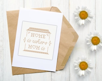 May include: A white card with a brown cross-stitch design that says "HOME is where MOM is". The card is sitting on a brown envelope with white daisies on a white wooden surface.