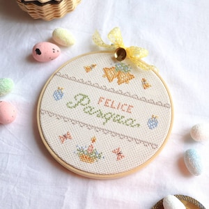 Può includere: Un cerchio da ricamo bianco con un disegno a punto croce che presenta le parole "FELICE PASQUA", che significa "Buona Pasqua". Il disegno include anche farfalle, pulcini e un cestino di uova. Il cerchio è circondato da uova di Pasqua color pastello.