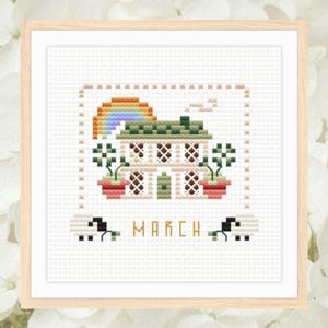 Pode incluir: Uma bordada em ponto cruz emoldurada que retrata uma casa com um arco-íris, plantas em vasos e a palavra "MARÇO". A casa tem um telhado verde e paredes brancas. O arco-íris arqueia-se sobre a casa, e as plantas estão em vasos vermelhos.