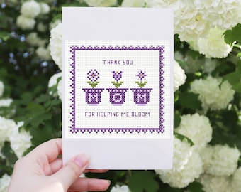 May include: A white card with a purple border and a cross-stitch design. The design features three purple flower pots with the word "MOM" spelled out in the pots. The text reads "Thank you for helping me bloom".