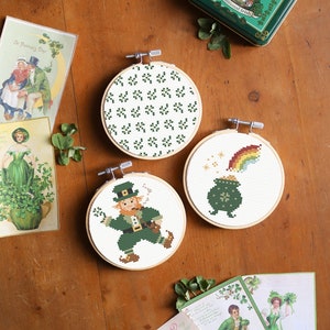 Puede incluir: Tres diseños de bordado de punto de cruz enmarcados con un tema del Día de San Patricio. Los diseños incluyen una olla de oro al final de un arcoíris, un duende y un patrón de tréboles.