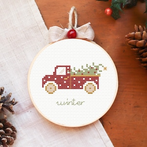 May include: A red truck with white polka dots carrying a green Christmas tree. The truck is embroidered on a white fabric circle with a wooden frame. The word "winter" is embroidered below the truck.
