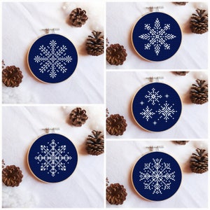 May include: Four embroidery hoops with navy blue fabric and white snowflake designs. The snowflakes are different designs, including a single snowflake, a snowflake with smaller snowflakes around it, and two different single snowflakes.