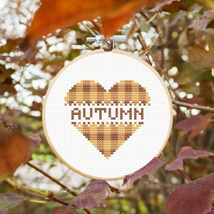 Peut inclure: Un cœur en tissu à carreaux bruns et beiges avec le mot "AUTUMN" brodé à l'intérieur. Le cœur est dans un cercle de broderie blanc, suspendu à une branche avec des feuilles brunes.