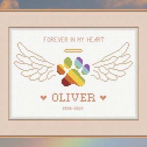 May include: A white cross-stitch embroidery with a rainbow paw print, angel wings, and the text "FOREVER IN MY HEART" above the paw print. Below the paw print is the name "OLIVER" and the dates "2008-2022".