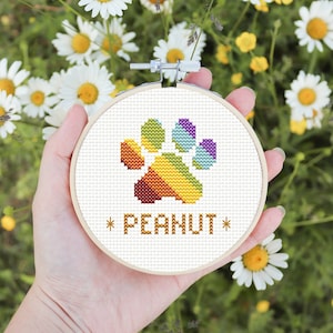 Peut inclure: Un cercle de broderie blanc avec une empreinte de patte arc-en-ciel et le texte "*PEANUT*" en fil doré. Le cercle est tenu dans une main sur un fond de marguerites blanches.