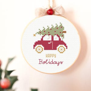 May include: A cross-stitch embroidery hoop featuring a red car carrying a green Christmas tree. The text "HAPPY Holidays" is stitched below the car. The hoop is adorned with a beige bow and a red berry.