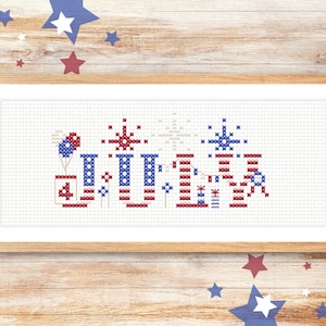 May include: A framed cross-stitch piece featuring a patriotic design. The artwork showcases red, white, and blue elements, including fireworks, balloons, and the number "4". The piece is framed in a light-colored wooden frame.