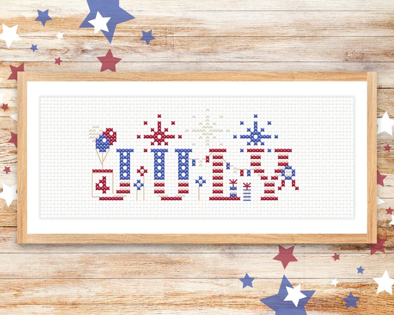 May include: A framed cross-stitch piece featuring a patriotic design. The artwork showcases red, white, and blue elements, including fireworks, balloons, and the number "4". The piece is framed in a light-colored wooden frame.