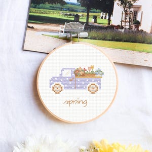 May include: A purple and white polka dot truck with a bed full of colorful flowers. The truck is embroidered on a white canvas and framed in a wooden hoop. The word "spring" is embroidered below the truck.