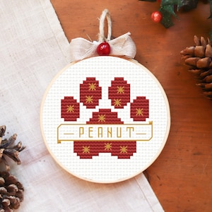 May include: A round, cross-stitched ornament featuring a paw print design in shades of red and gold on a white background. The name "PEANUT" is stitched below the paw print. The ornament is trimmed with a beige ribbon and a red berry.