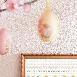 May include: A framed cross-stitch design featuring a brown bunny rabbit surrounded by yellow and green Easter eggs. The design is on white fabric and has a white border.