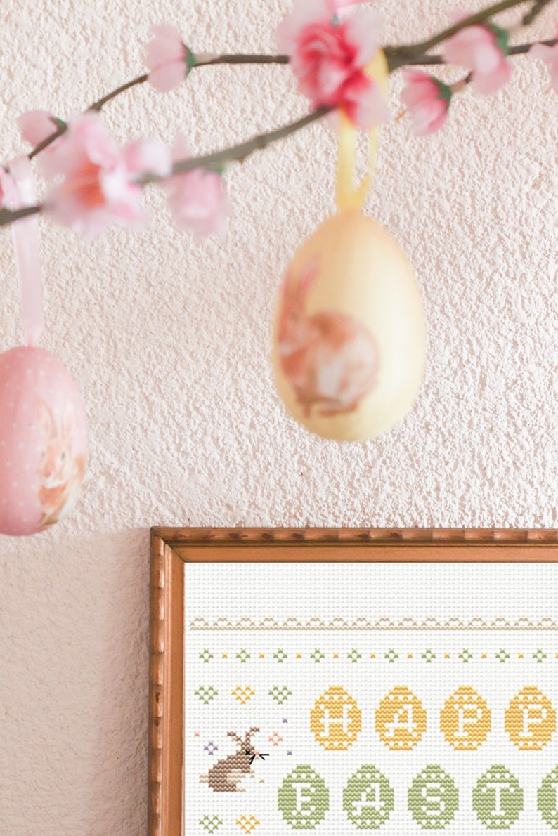 May include: A framed cross-stitch design featuring a brown bunny rabbit surrounded by yellow and green Easter eggs. The design is on white fabric and has a white border.