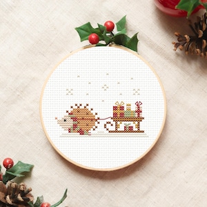 Può includere: Un telaio da ricamo a punto croce con un riccio che tira una slitta piena di regali. Il disegno utilizza fili marroni, rossi e verdi su sfondo bianco. Il telaio è incorniciato in legno naturale, con agrifoglio e bacche in alto.