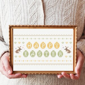 May include: A framed cross-stitch design featuring the words "Happy Easter" surrounded by yellow and green eggs and two brown bunnies.
