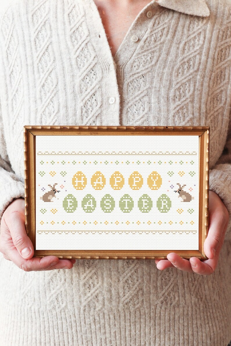 May include: A framed cross-stitch design featuring the words "Happy Easter" surrounded by yellow and green eggs and two brown bunnies.
