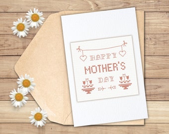 Puede incluir: Una tarjeta blanca con un mensaje bordado en rojo que dice "Feliz Día de la Madre". La tarjeta está decorada con corazones y flores.