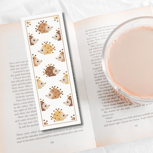 May include: A cross-stitch bookmark with a repeating pattern of hedgehogs in brown and yellow. The bookmark is rectangular and is placed on an open book, with a glass of pink liquid in the background. The bookmark is approximately 18 cm long.
