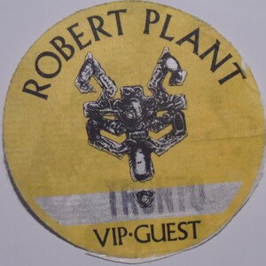 May include: A round, yellow VIP guest pass with the text "ROBERT PLANT" in black, arched around the top. A black and white graphic is in the center. The bottom of the pass reads "VIP GUEST".