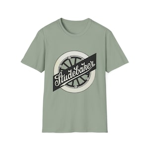 May include: A sage green short-sleeve t-shirt featuring a central graphic of a black and white vintage spoked wheel. A black diagonal banner overlays the wheel, displaying the word Studebaker in a white, cursive, stylized script font.