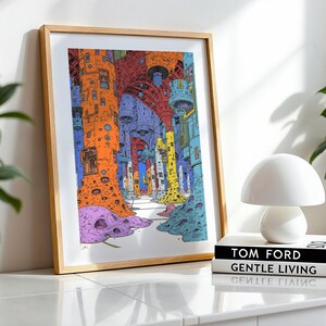 May include: A framed print of a colorful, abstract illustration depicting a city with tall, multi-colored buildings. The print is in a light wood frame and is sitting on a white table with a white lamp and books.