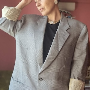 May include: A gray plaid blazer with a notched lapel and two front pockets. The blazer has a classic fit and is worn with a black top. The sleeves are cuffed to reveal a cream-colored lining. The blazer is part of a vintage clothing collection.