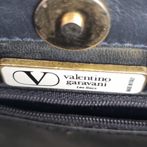 May include: Close-up of a black leather handbag with a gold-toned clasp. The handbag features a rectangular tag with the Valentino Garavani logo and the text "Les Sacs". The tag also reads "Made in Italy".