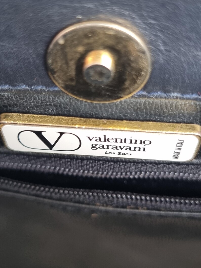 May include: Close-up of a black leather handbag with a gold-toned clasp. The handbag features a rectangular tag with the Valentino Garavani logo and the text "Les Sacs". The tag also reads "Made in Italy".
