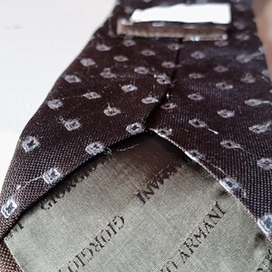 May include: A dark brown silk tie with a repeating white geometric pattern. The tie is labeled "GIORGIO ARMANI" in white text on a green fabric. The tie is a classic accessory for formal or business attire.