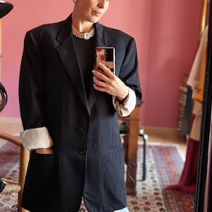 May include: A dark navy blazer with rolled-up sleeves revealing a white lining. The blazer is paired with light blue jeans. A gold necklace with a large pendant is visible. The person is holding a phone with a patterned case.